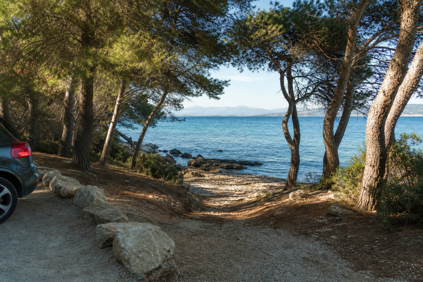 KI-generiertes Stimmungsbild eines einfachen Zugangs durch schattige Pinien zu einem steinigen Strand mit klarem Meer