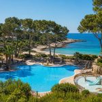 KI-generiertes Stimmungsbild mit Familienpool, Kinderwasserbereich, Pinien und Mittelmeerbucht für das Iberostar Waves Pinos Park in Font de sa Cala